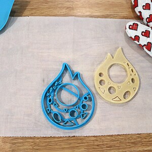 May include: Two blue and beige plastic cookie cutters shaped like flames with intricate designs. The cutters are on a white parchment paper surface.