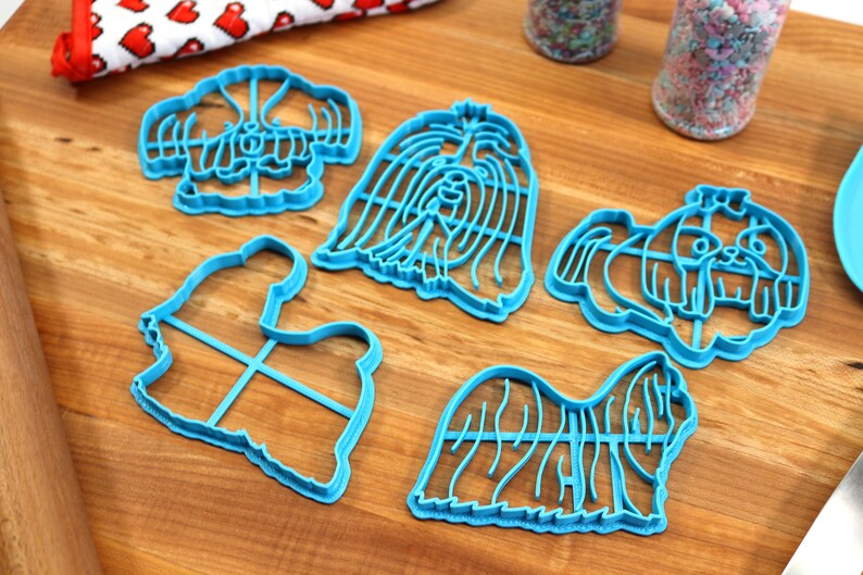 May include: Five blue plastic cookie cutters shaped like Shih Tzu dogs. The cutters are on a wooden surface.