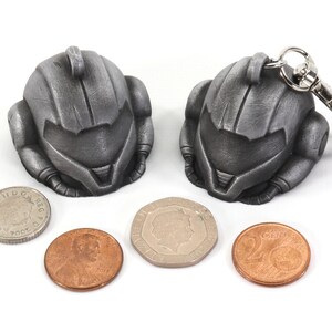 Puede incluir: Dos llaveros de metal de color plateado con forma de casco. Los llaveros est&aacute;n al lado de tres monedas, una plateada y dos de cobre.
