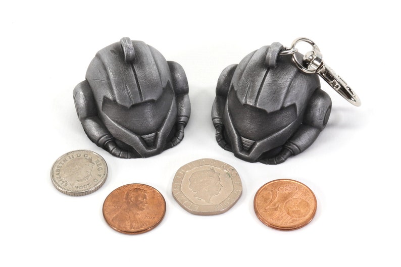 Puede incluir: Dos llaveros de metal de color plateado con forma de casco. Los llaveros est&aacute;n al lado de tres monedas, una plateada y dos de cobre.