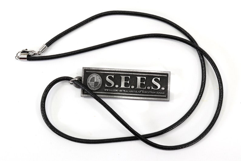 May include: A black cord necklace with a silver pendant. The pendant is rectangular and has the text "S.E.E.S. SPECIALIZED EXTRACURRICULAR EXECUTION SQUAD" on it.