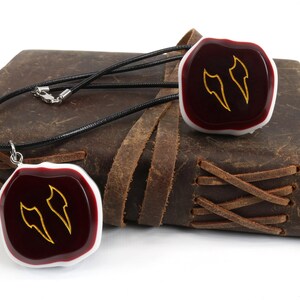 Op de afbeelding: Een ketting van zwart lederen koord met twee rode en witte hars hangers. Elke hanger heeft een gouden ontwerp van twee gestileerde vleugels.