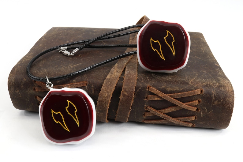 Op de afbeelding: Een ketting van zwart lederen koord met twee rode en witte hars hangers. Elke hanger heeft een gouden ontwerp van twee gestileerde vleugels.