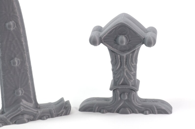 May include: A gray 3D printed model of a Viking axe head with a detailed handle and a decorative top. The axe head is designed with a textured surface and a raised design on the top.
