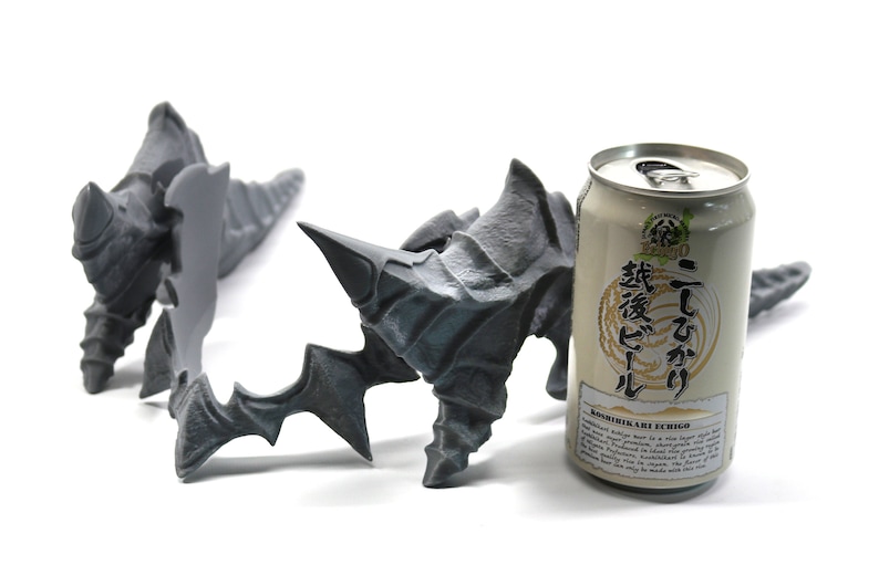 May include: Two grey 3D printed sculptures resembling claws or horns, with a can of Japanese beer in the background. The can is white with black text that reads "Koshihikari ECHIGO" and "越後こしひかり" in Japanese.
