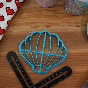 May include: A blue plastic seashell-shaped cookie cutter on a wooden surface. The cutter has a detailed design with multiple ridges.