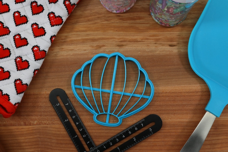 May include: A blue plastic seashell-shaped cookie cutter on a wooden surface. The cutter has a detailed design with multiple ridges.