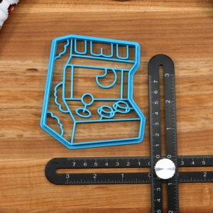 May include: A blue plastic cookie cutter shaped like an arcade game. The cutter has a retro design with a joystick, buttons, and a screen.