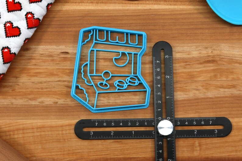 May include: A blue plastic cookie cutter shaped like an arcade game. The cutter has a retro design with a joystick, buttons, and a screen.