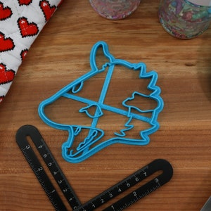 May include: A blue plastic cookie cutter shaped like a wolf's head. The cutter is on a wooden surface with a ruler and a blue spatula.