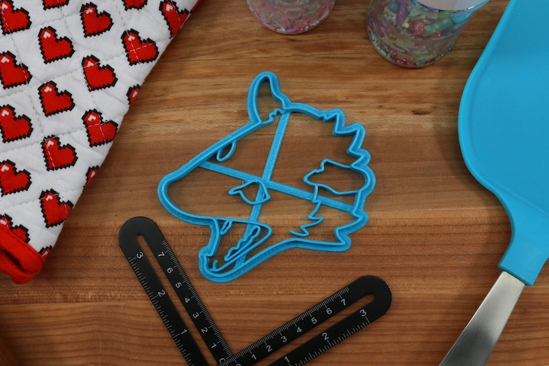 May include: A blue plastic cookie cutter shaped like a wolf's head. The cutter is on a wooden surface with a ruler and a blue spatula.