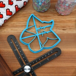 May include: A blue plastic cookie cutter shaped like a star with a decorative design. The cutter is on a wooden surface with a ruler and a rolling pin.