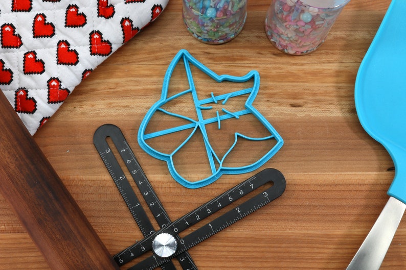 May include: A blue plastic cookie cutter shaped like a star with a decorative design. The cutter is on a wooden surface with a ruler and a rolling pin.