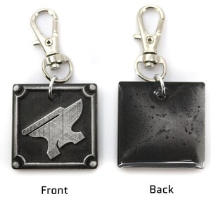 May include: A silver metal keychain with a square charm. The front of the charm features a silver anvil design. The back of the charm is plain and silver.