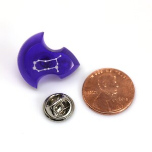 May include: A purple resin pin with a white outline of the Gemini constellation. The pin is next to a US penny for size comparison.