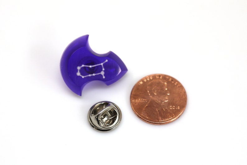May include: A purple resin pin with a white outline of the Gemini constellation. The pin is next to a US penny for size comparison.
