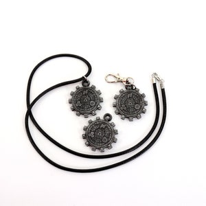 May include: Three silver-toned metal pendants with intricate designs, two on black cords and one on a silver chain. The pendants feature a circular shape with a gear-like design and a central symbol.