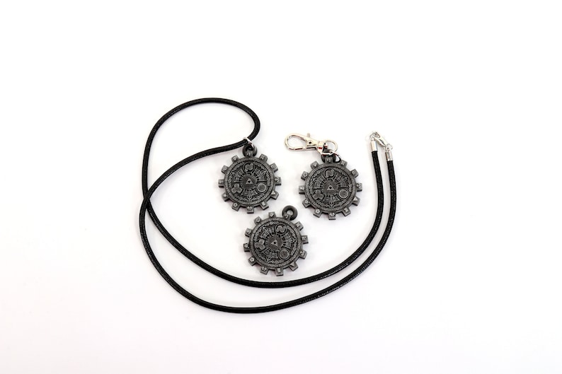 May include: Three silver-toned metal pendants with intricate designs, two on black cords and one on a silver chain. The pendants feature a circular shape with a gear-like design and a central symbol.