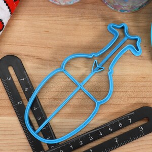 May include: A blue plastic cookie cutter shaped like a violin. The cutter is approximately 10 cm long and 5 cm wide.