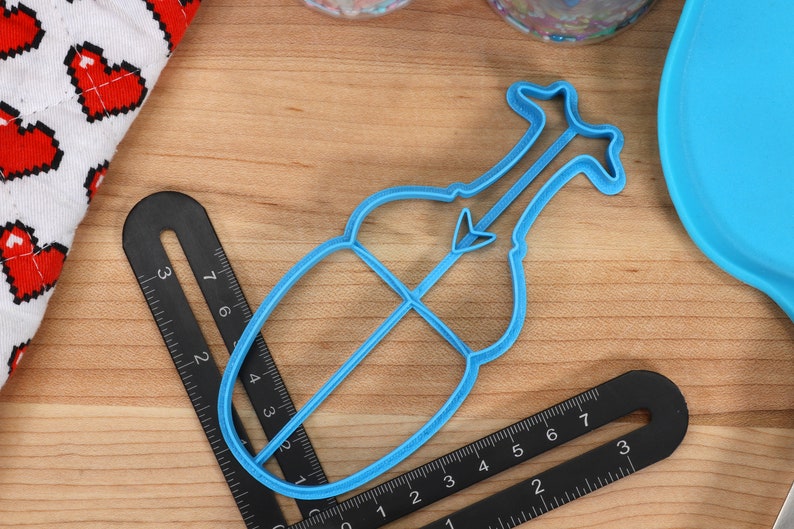 May include: A blue plastic cookie cutter shaped like a violin. The cutter is approximately 10 cm long and 5 cm wide.