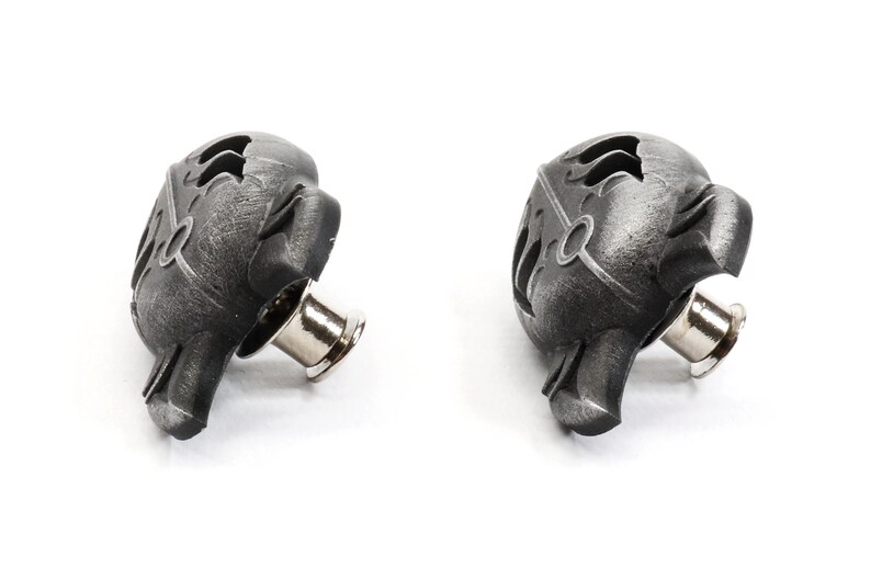 Op de afbeelding: Twee zwarte metalen drukknopen met een decoratief ontwerp. De drukknopen hebben een zilveren metalen pen.
