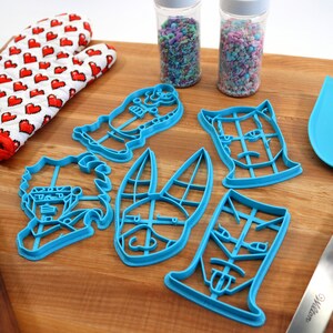May include: A set of six blue plastic cookie cutters shaped like cartoon faces. The cutters are on a wooden cutting board with a rolling pin, a pair of red oven mitts, and a spatula.