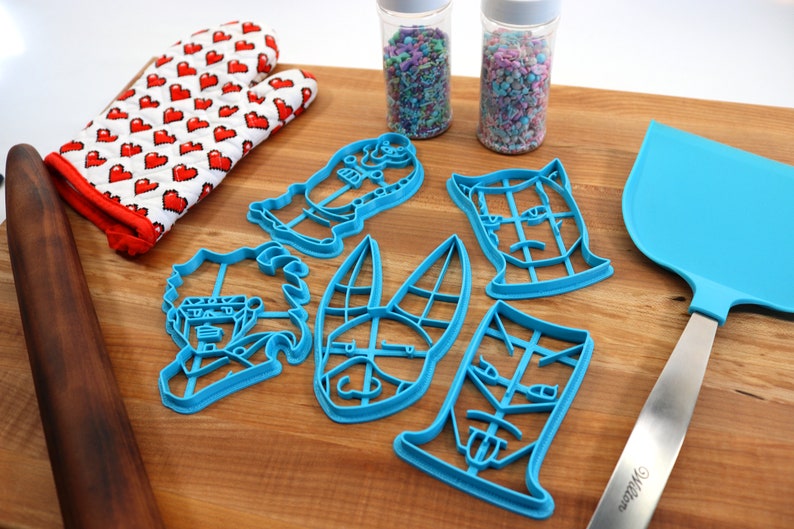 May include: A set of six blue plastic cookie cutters shaped like cartoon faces. The cutters are on a wooden cutting board with a rolling pin, a pair of red oven mitts, and a spatula.