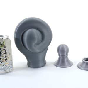 K&ouml;nnte beinhalten: Eine graue, 3D-gedruckte Skulptur mit Spiraldesign, neben zwei kleineren, passenden grauen Objekten und einer Dose japanischen Biers. Die Objekte befinden sich auf einer wei&szlig;en Oberfl&auml;che.
