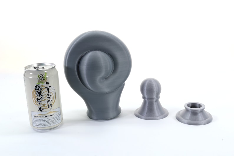 K&ouml;nnte beinhalten: Eine graue, 3D-gedruckte Skulptur mit Spiraldesign, neben zwei kleineren, passenden grauen Objekten und einer Dose japanischen Biers. Die Objekte befinden sich auf einer wei&szlig;en Oberfl&auml;che.