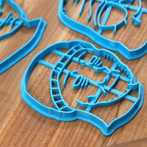 May include: Three blue plastic cookie cutters shaped like faces. The cutters are on a wooden surface.