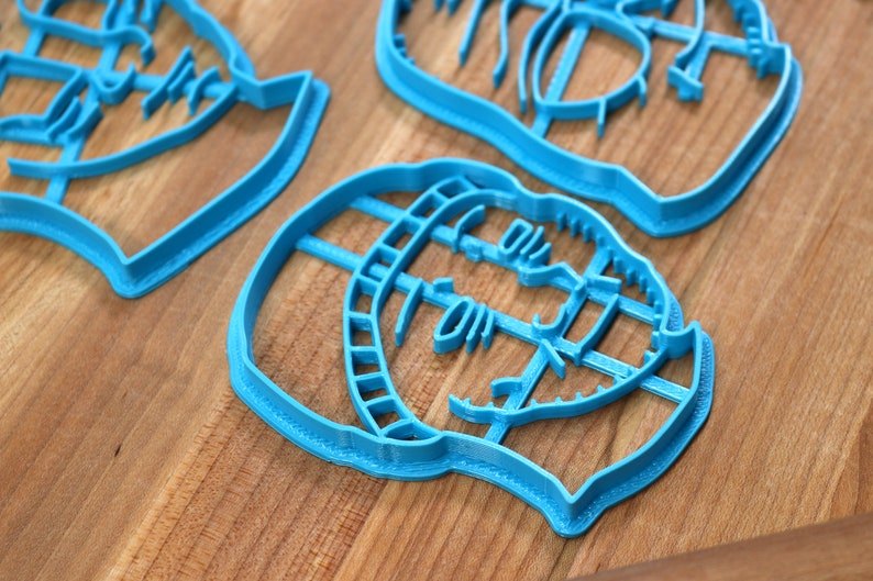 May include: Three blue plastic cookie cutters shaped like faces. The cutters are on a wooden surface.