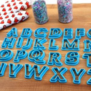 May include: A set of 26 blue plastic alphabet cookie cutters on a wooden surface. The cutters are arranged in alphabetical order.