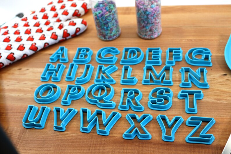 May include: A set of 26 blue plastic alphabet cookie cutters on a wooden surface. The cutters are arranged in alphabetical order.