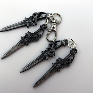 May include: Four silver-colored dagger keychains with intricate designs. Each dagger features a detailed hilt and blade, connected to a keyring and a clasp. The keychains are arranged on a white surface.