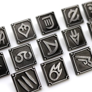 May include: Close-up of several square, dark metal pins with various symbols and designs. Each pin has a raised border and a detailed, embossed symbol in the center. The pins are arranged diagonally on a white surface.