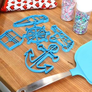 May include: A collection of blue plastic nautical-themed cookie cutters, including an anchor, ship's wheel, and sailboat, arranged on a wooden surface. Also present are a blue spatula, rolling pin, sprinkles, and a red and white heart-patterned oven mitt.