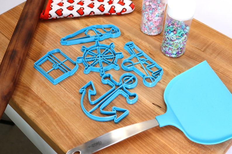 May include: A collection of blue plastic nautical-themed cookie cutters, including an anchor, ship's wheel, and sailboat, arranged on a wooden surface. Also present are a blue spatula, rolling pin, sprinkles, and a red and white heart-patterned oven mitt.