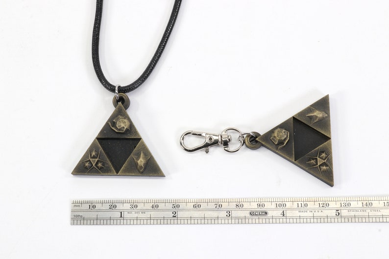 May include: A gold-colored pendant necklace and keychain featuring a triangular design with four symbols. The necklace has a black cord and the keychain has a silver metal clasp.