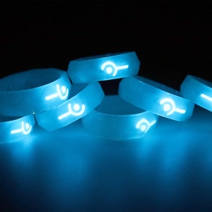May include: A group of six glow-in-the-dark silicone rings with a white symbol on each ring. The rings are stacked on top of each other and are glowing a bright blue color.