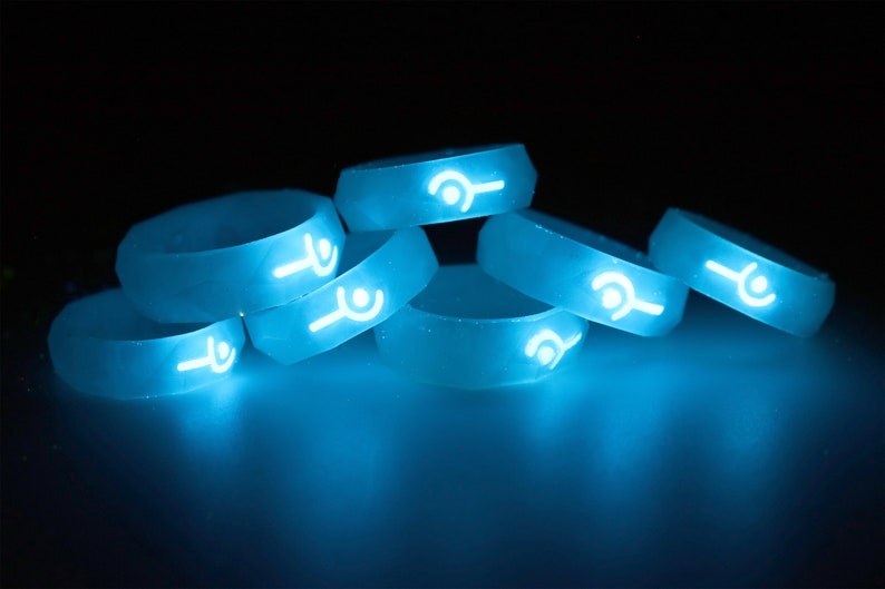May include: A group of six glow-in-the-dark silicone rings with a white symbol on each ring. The rings are stacked on top of each other and are glowing a bright blue color.
