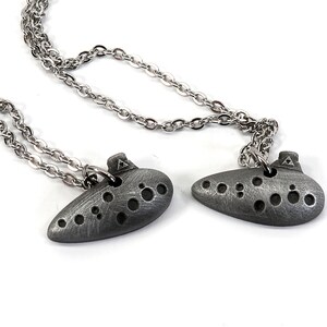 May include: Two silver ocarina necklaces with a silver chain. The ocarinas have a textured surface and are shaped like a traditional ocarina.