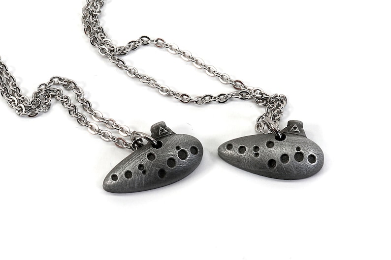 May include: Two silver ocarina necklaces with a silver chain. The ocarinas have a textured surface and are shaped like a traditional ocarina.