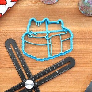 May include: Blue plastic cookie cutter shaped like two cats cuddling. The cutter is on a wooden surface.