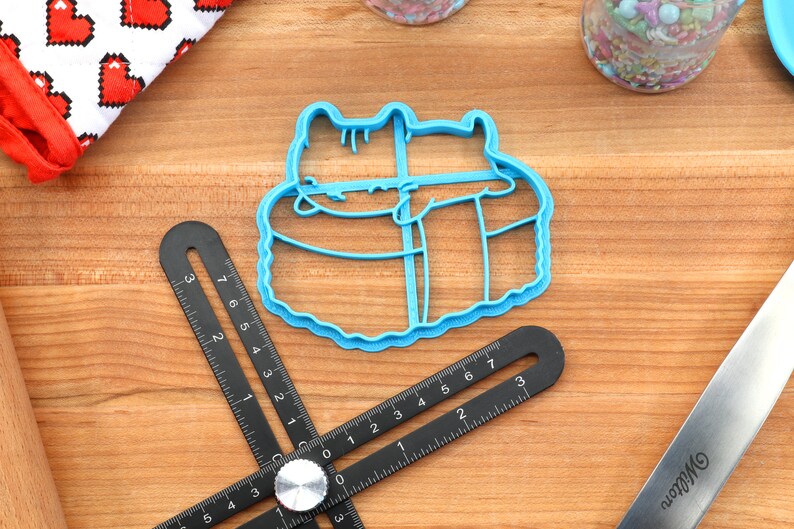 May include: Blue plastic cookie cutter shaped like two cats cuddling. The cutter is on a wooden surface.