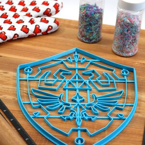 May include: A blue plastic cookie cutter shaped like the Triforce symbol from the Legend of Zelda video game series. The cookie cutter is on a wooden cutting board with a rolling pin, a ruler, and two jars of sprinkles.
