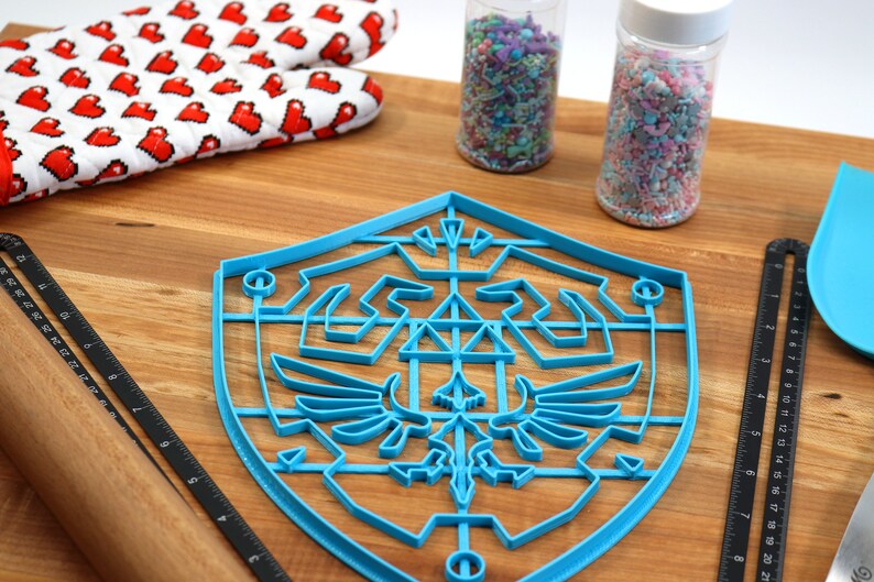 May include: A blue plastic cookie cutter shaped like the Triforce symbol from the Legend of Zelda video game series. The cookie cutter is on a wooden cutting board with a rolling pin, a ruler, and two jars of sprinkles.