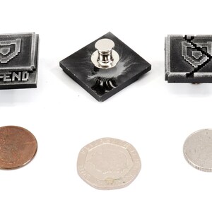 May include: Three silver-colored metal pins with a pixelated shield design. The pins are square shaped and have a black background. The text "DEFEND" is printed on one of the pins. The pins are shown next to three coins.