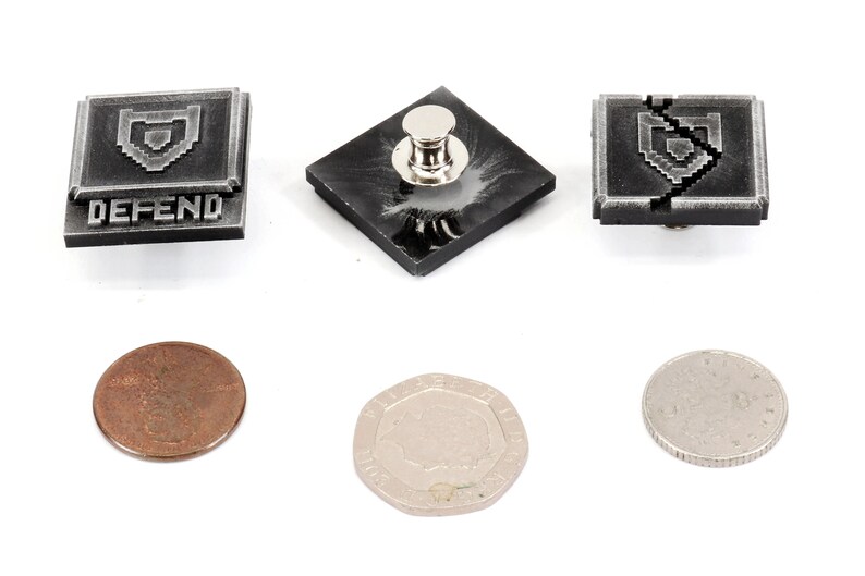 May include: Three silver-colored metal pins with a pixelated shield design. The pins are square shaped and have a black background. The text "DEFEND" is printed on one of the pins. The pins are shown next to three coins.