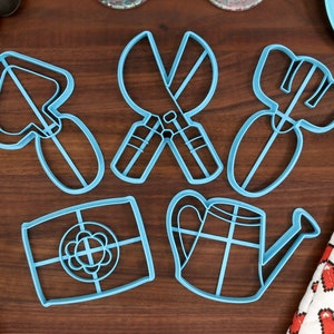 May include: A set of six blue plastic cookie cutters shaped like gardening tools. The cutters include a trowel, a pair of shears, a rake, a watering can, a flower, and a square.