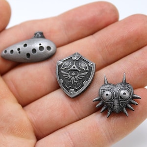 May include: Three silver-coloured metal pins featuring iconic symbols from the Legend of Zelda video game series. The pins depict the Ocarina of Time, the Hylian Shield, and Majora's Mask.